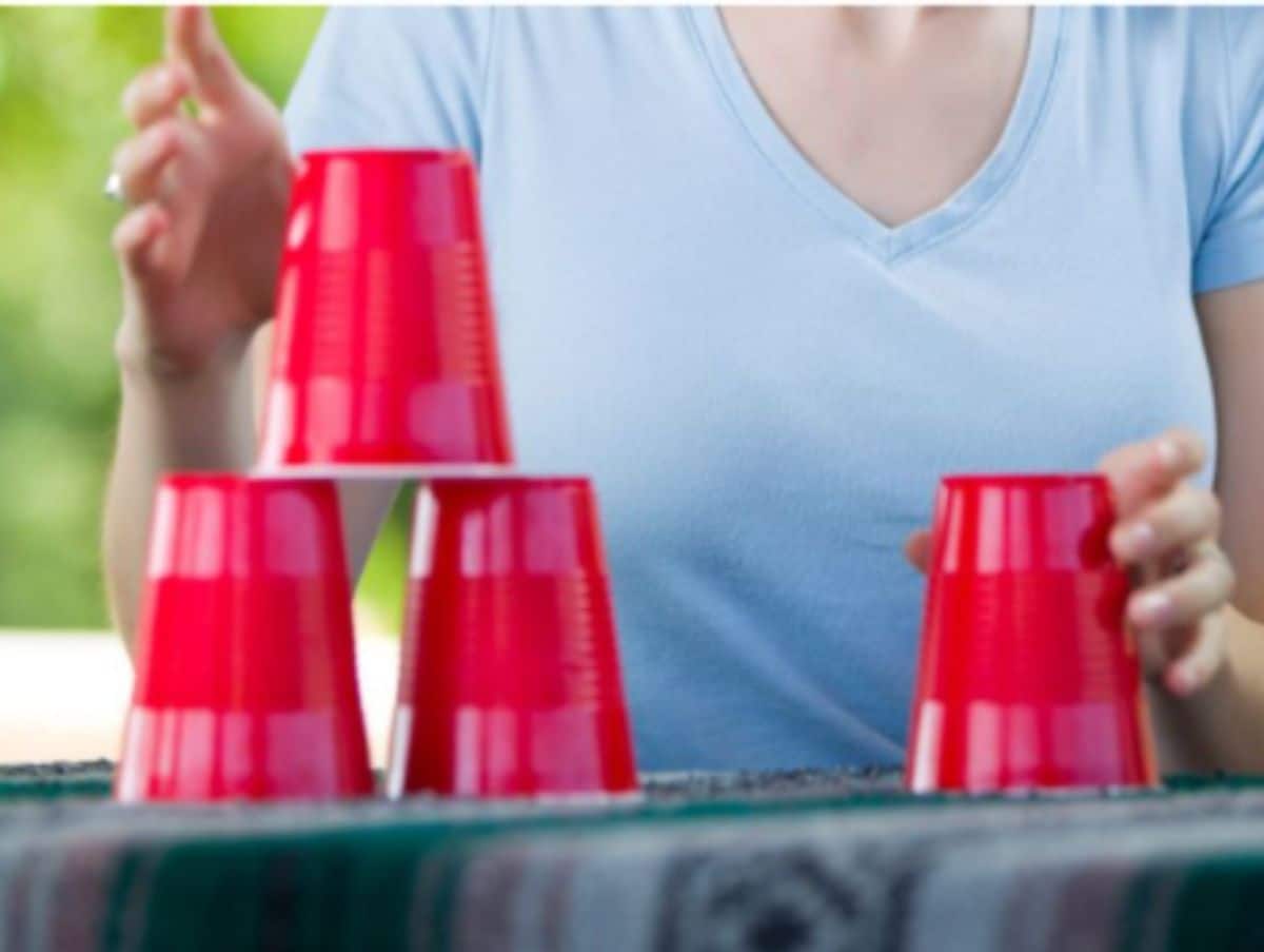 a woman in a blue shirt sits behind an outdoor table. She is stacking 4 red solo cups on the table.