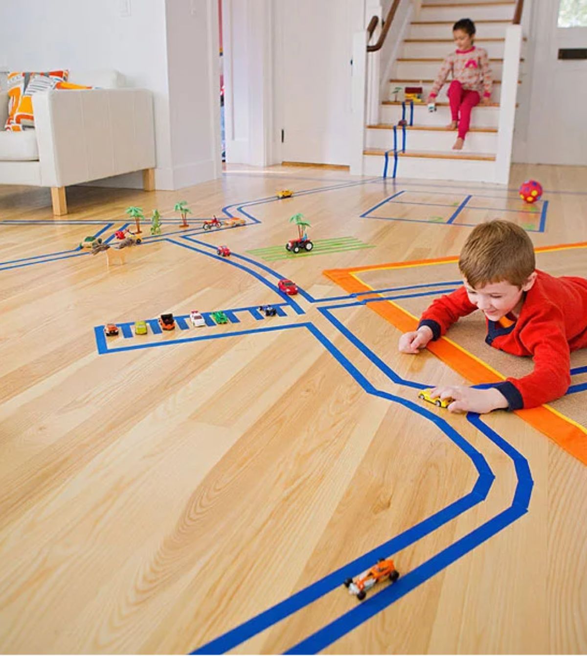 a pale laminate floor is covered in tracks made from colored tape. 2 children are pushing cars along the tracks