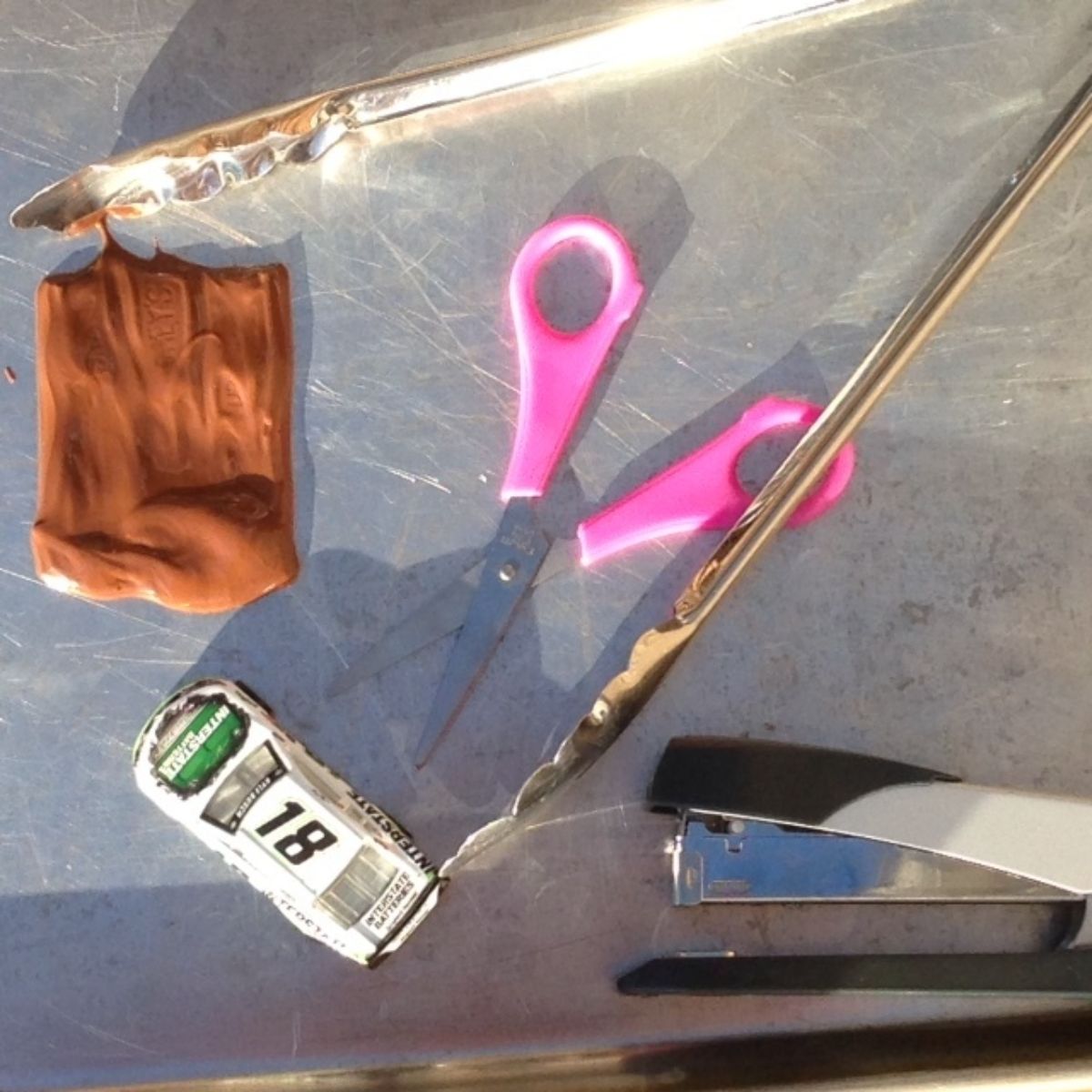 on a metal top is a collection of items: a pair of pink scissors, chocolate, metal tongs, a toy car and a stapler