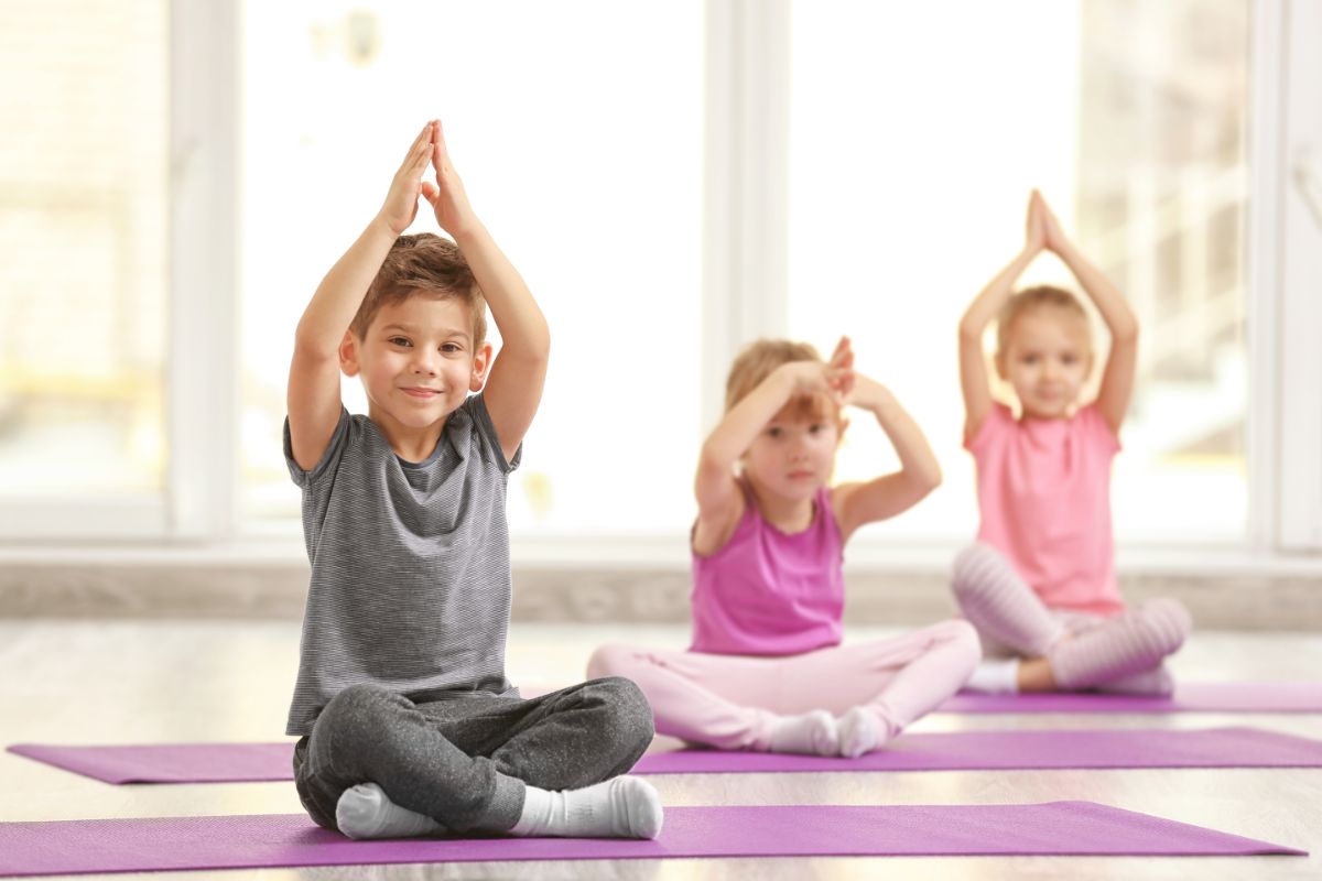 3 children sit on purple yoga mats cross liegged with their hands in prayer pose