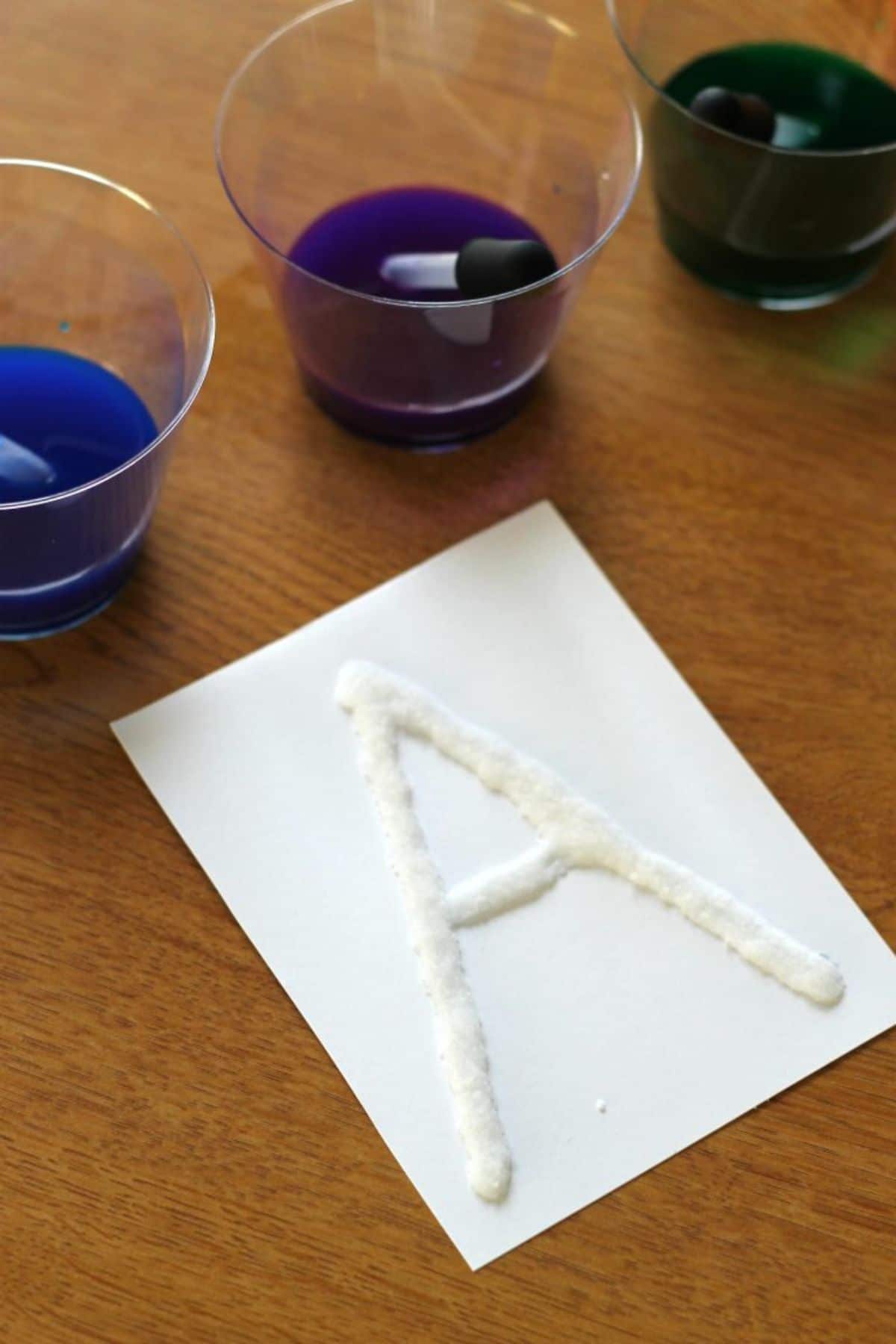 on a table is a sheet of paper with an "A" written in salt. Behind the sheet are 3 glasses filled with coloured water