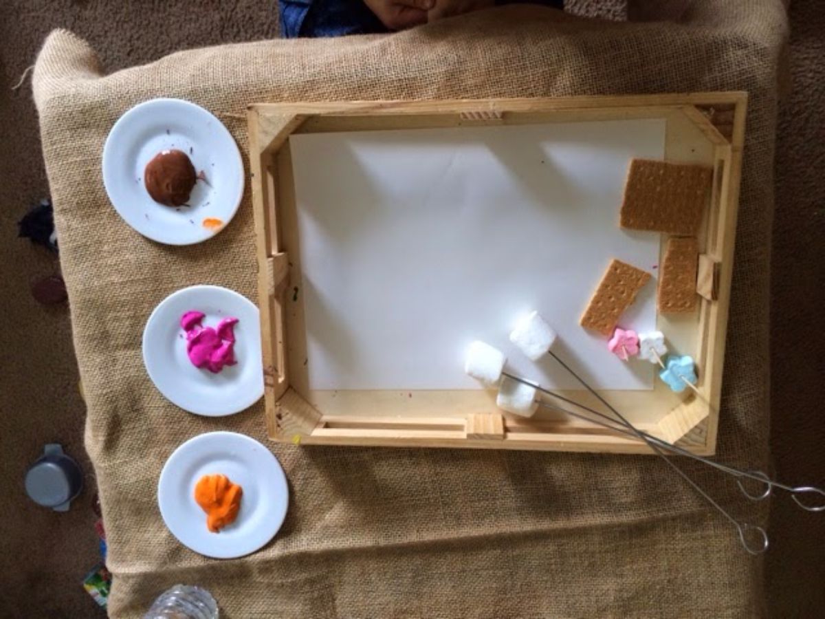 on a hessian mat is a tray with paper, marshmallows on skewers and gram crackers. To the side are 3 saucers with brown, pink and orange paint on them