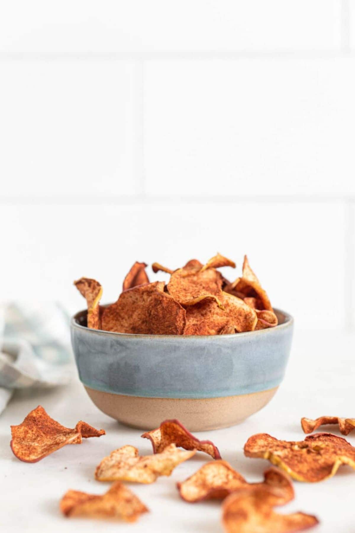a ceramic bowl of baked chips. apple chips are scattered around the bowl