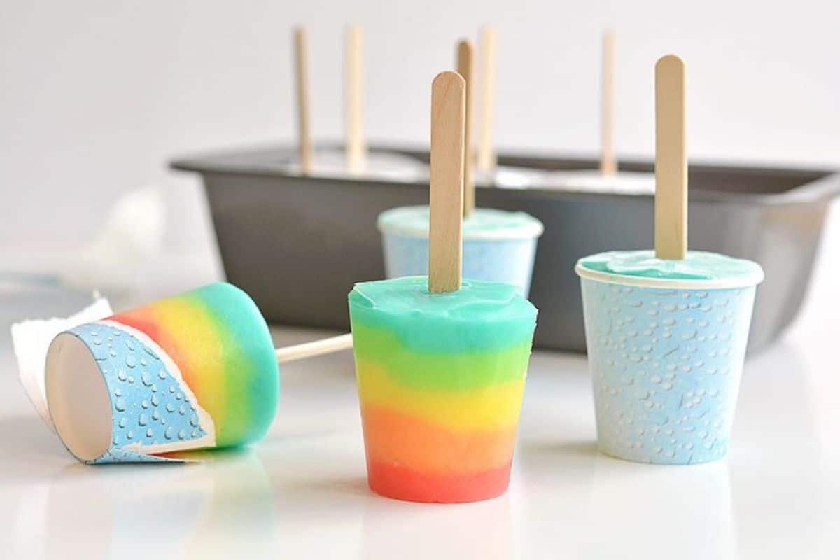 4 rainbow popsicles are in the process of being taken out of the paper cups. Behind is a loaf tin
