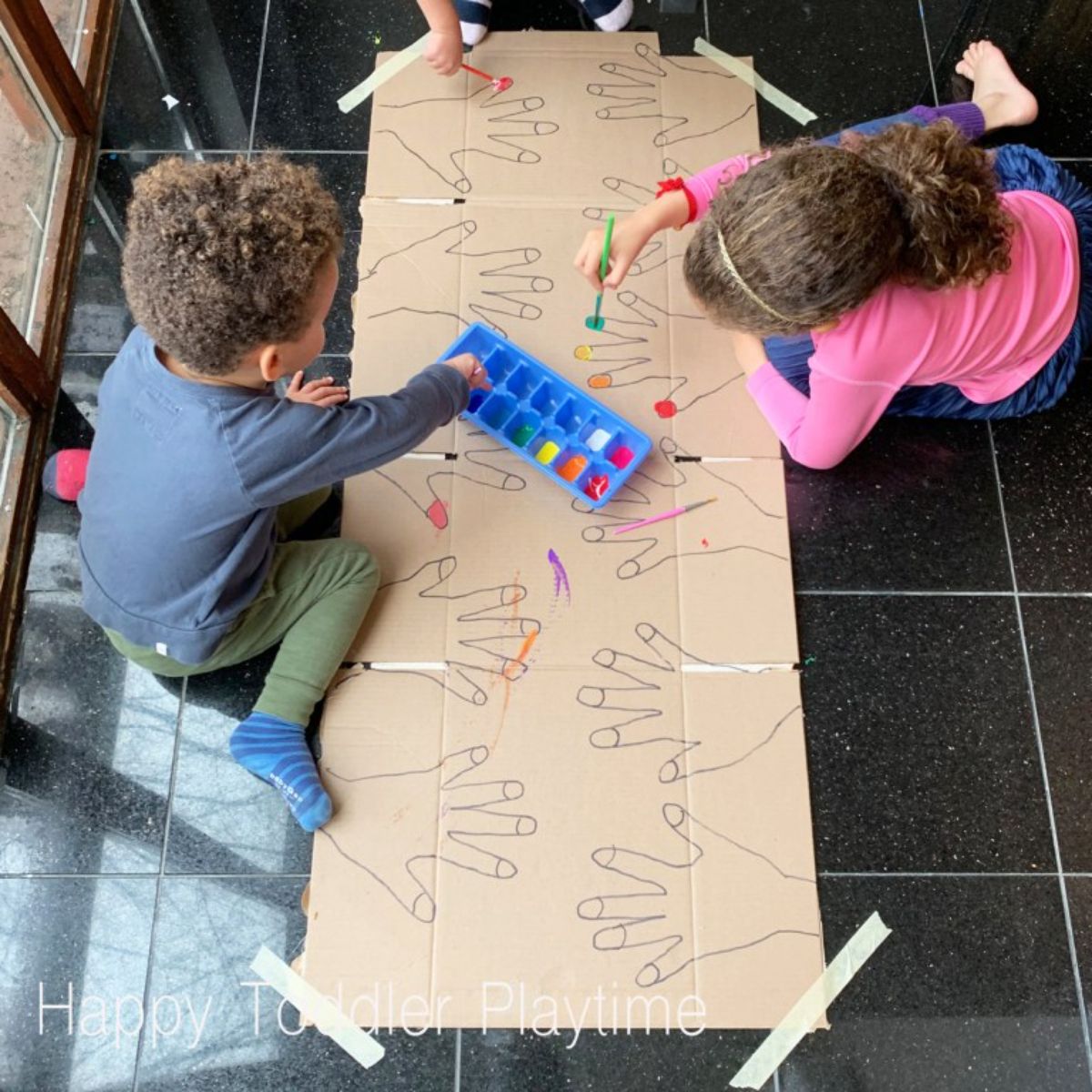 two children kneel on a black tiled floor coloring in large hand shapes drawn on a sheet of cardboard