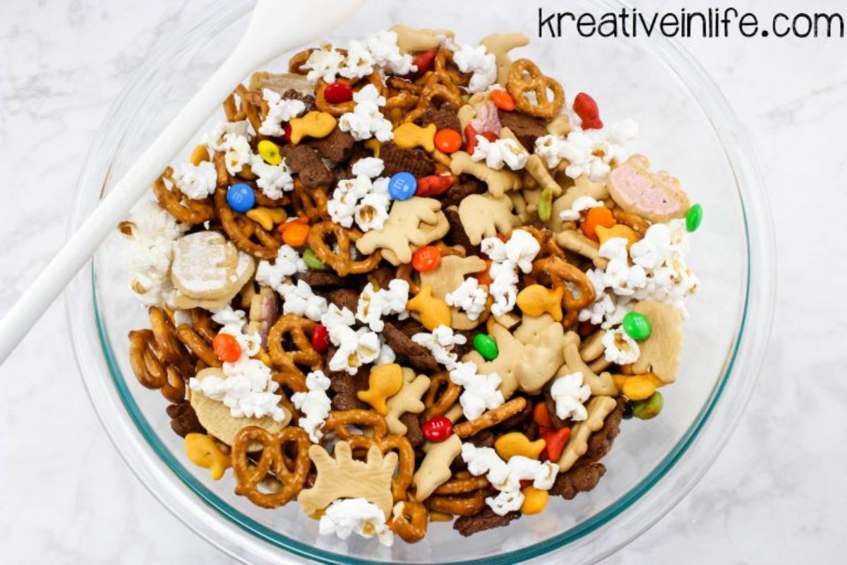 a pyrex bowl full of trail mix containing pretzels, animal crackers, candy and popcorn