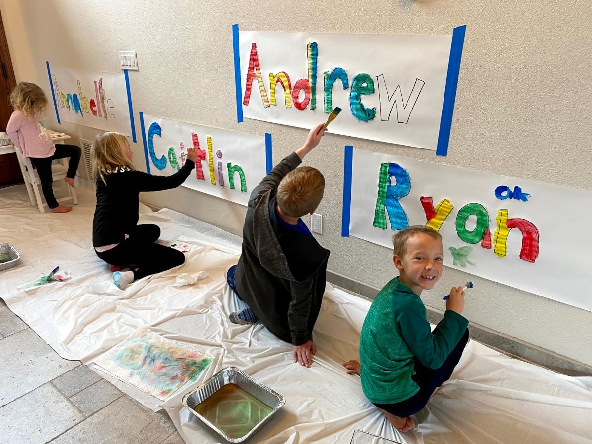 4 children are sitting on a sheet and painting their names on pieces of paper taped to the wall
