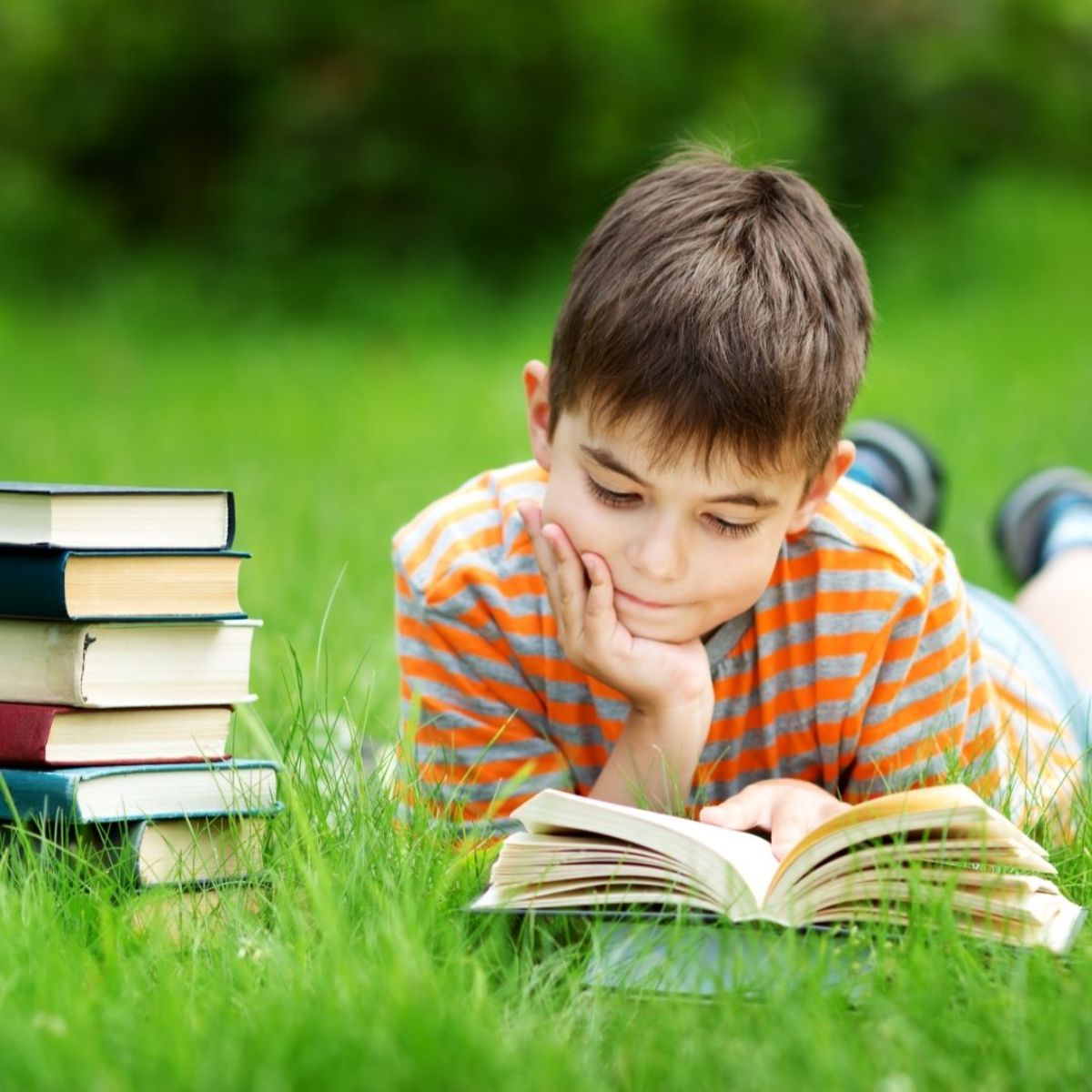 a boy in a ornage and grey striped shirt lays on the grass reading a book with a pile of books next to him