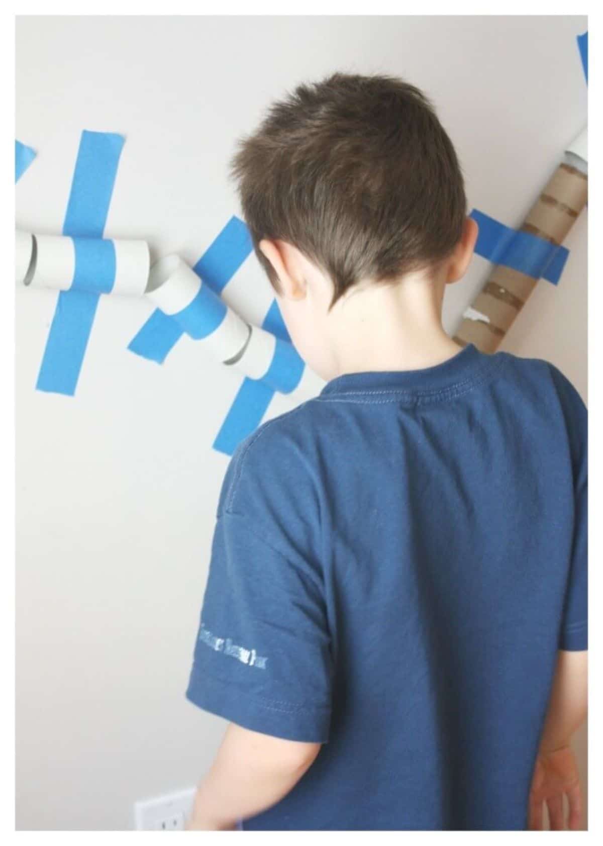 A boy in a blue shirt stands in front of a wall that has toilet roll tibes taped to it