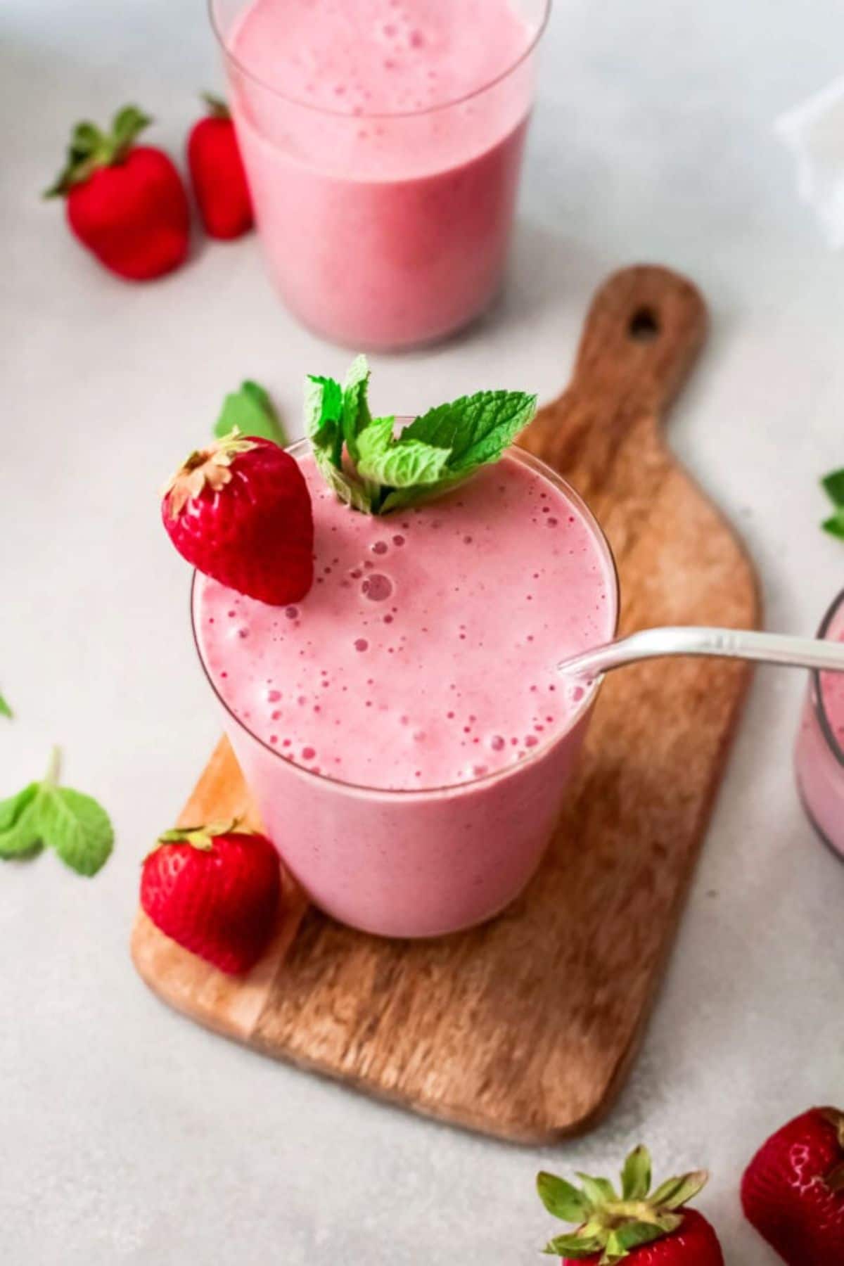 on a wooden board is a glass full of a pink smoothie, garnished with a strawberry and mint sprig.
