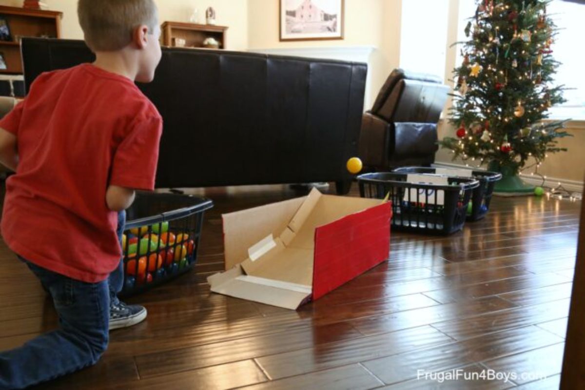 a boy kneels in a living room throwing a ping pong ball at a cardboard box. A christmas tree is in the background