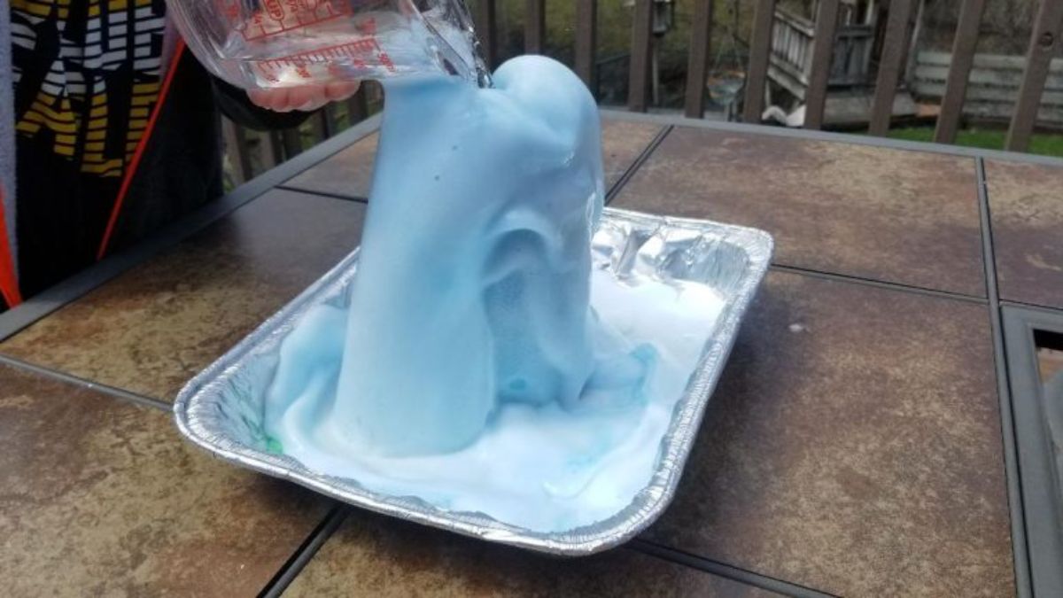 on an outdoor table is a tin tray. On the tray is a load of blue foam. A glass pyrex jug pours water onto it