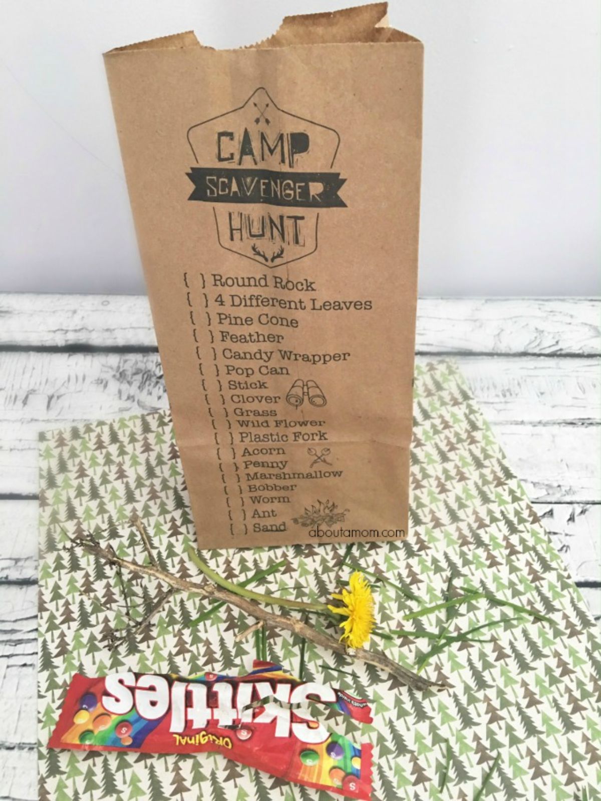 A white table with a piece of fabric laying on it printed with trees. Grass and flowers are scattered on top and a packet of skittles is in front. On top of the fabric is a brown paper bag with a scavenger hunt list printed onto it