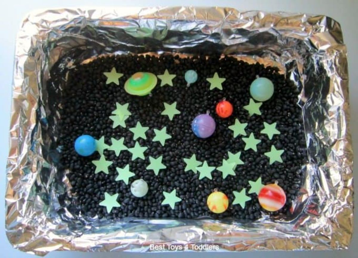 a deep tray is covered in silver foil. In it are black lentills, glow in the dark stars and plastic planets