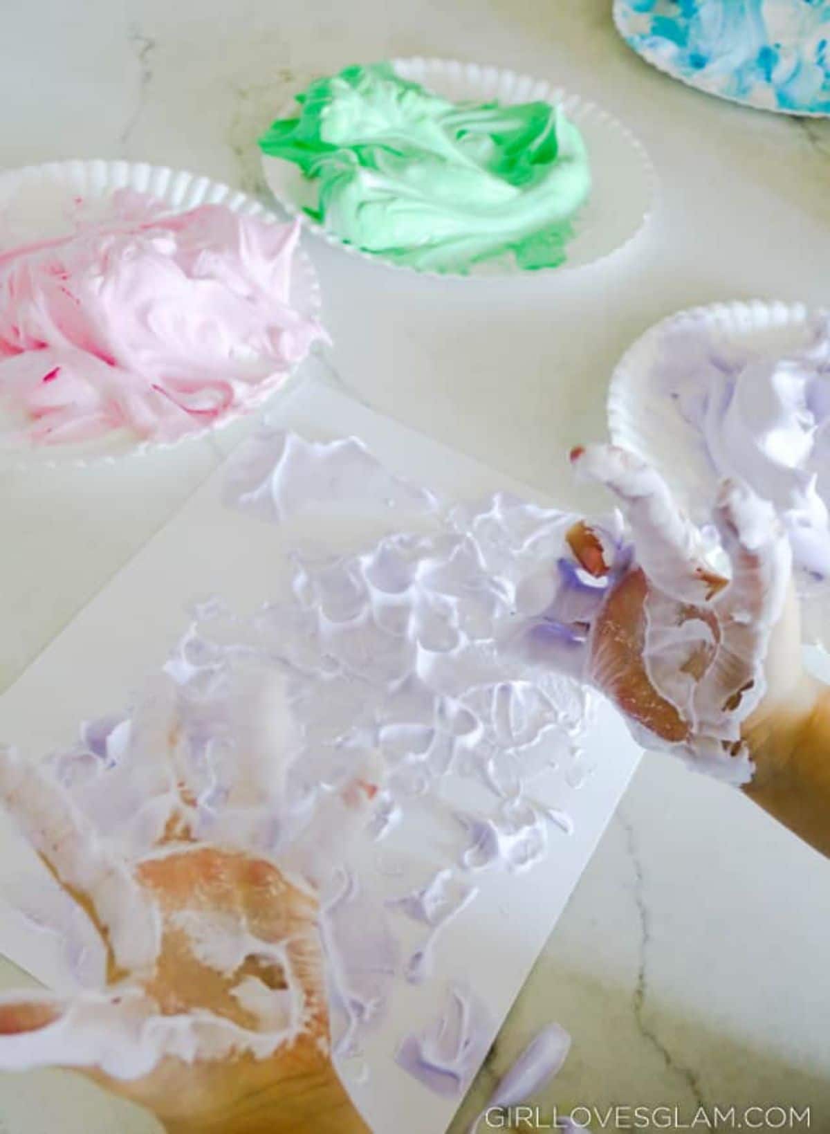 two hands are covered in foam. A sheet of paper is also covered in colored foam. 4 paper plates hold different colored foam