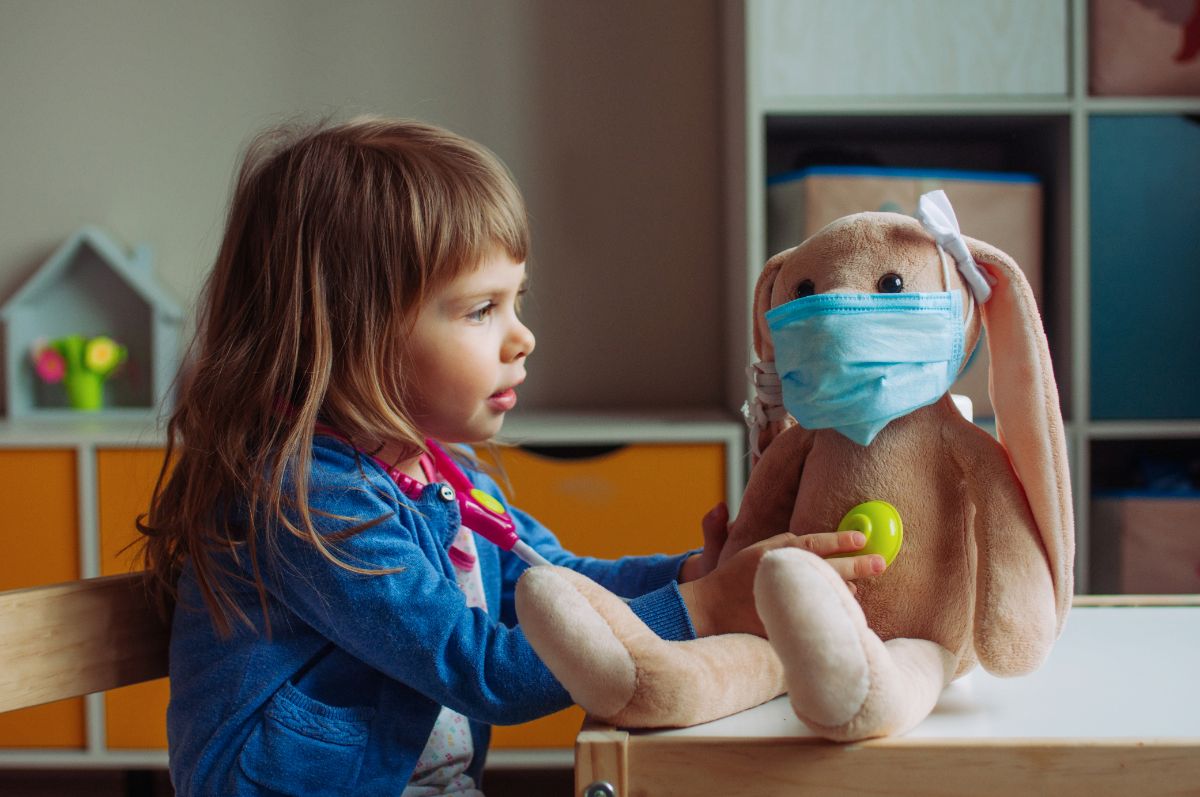 a girl in a blue shirt with long brown hair holds the end of a stethoscope up to the chest of a soft toy rabbit who is wearing a surgical mask