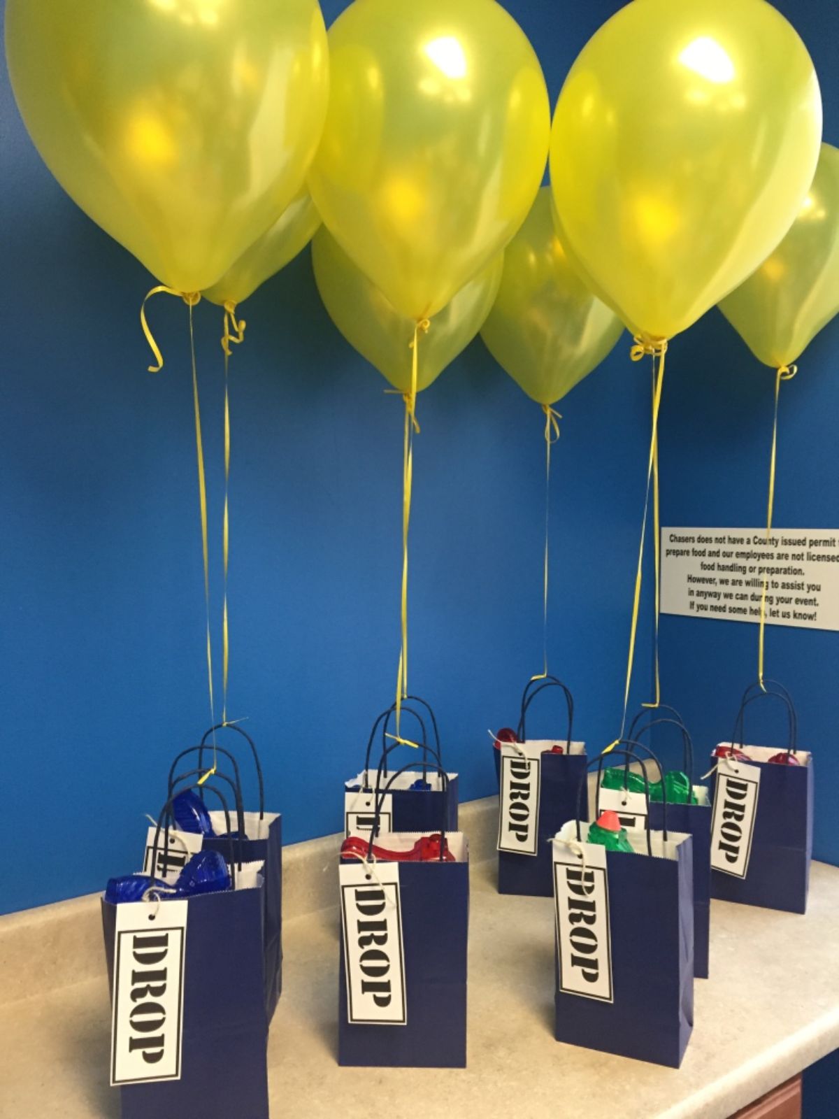 on a table, 8 blue bags labelled with "DROP" and attached to yellow balloons