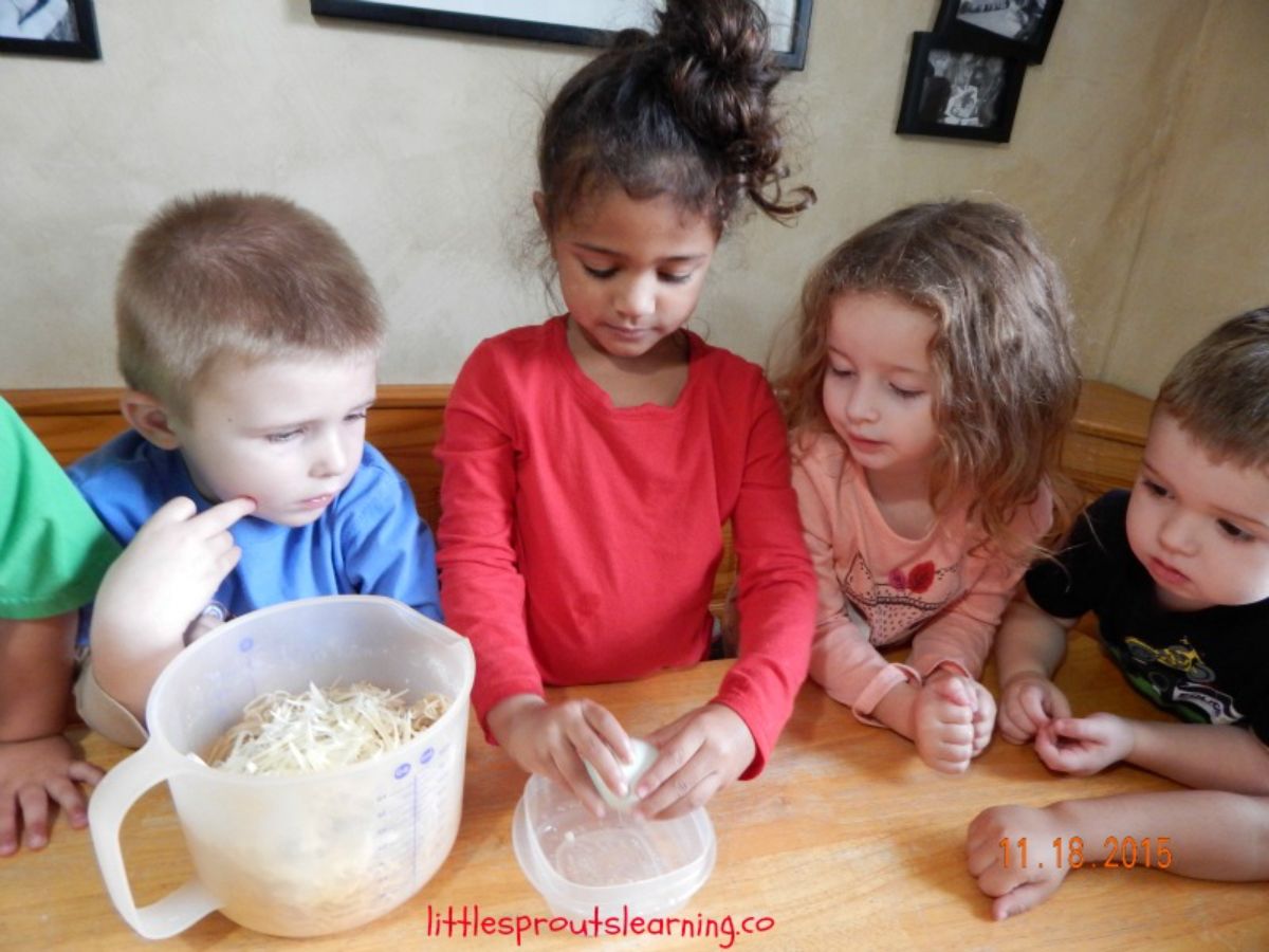 a group of children sit around a table. A black girl in a red shirt is holding a ball of mozzarella and pulling it apart while the other children watch. A jug full of cheese and egg is to the side