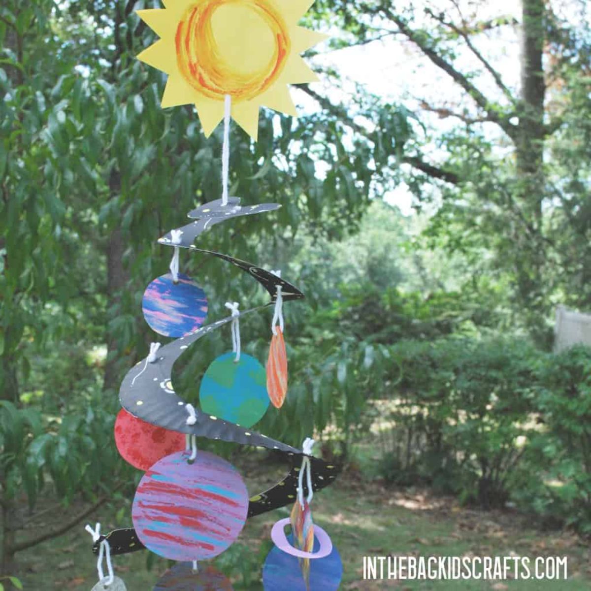 a swirled wind chime made of paper and decorated with the solar system hangs in front of trees