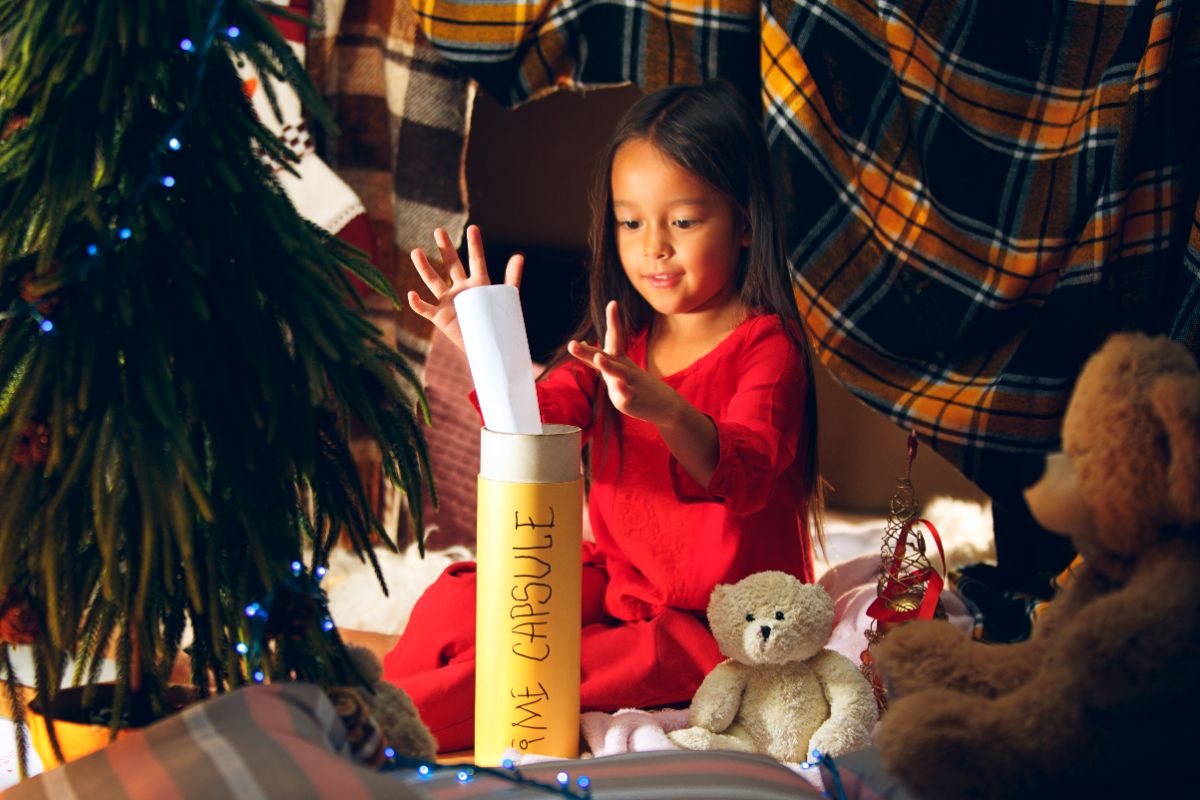 an asian girl in a red dress places a rolled up piece of paper into a cardboard tube labelled "Time Capsule" She is surrounded by a tartan sheet and soft toys