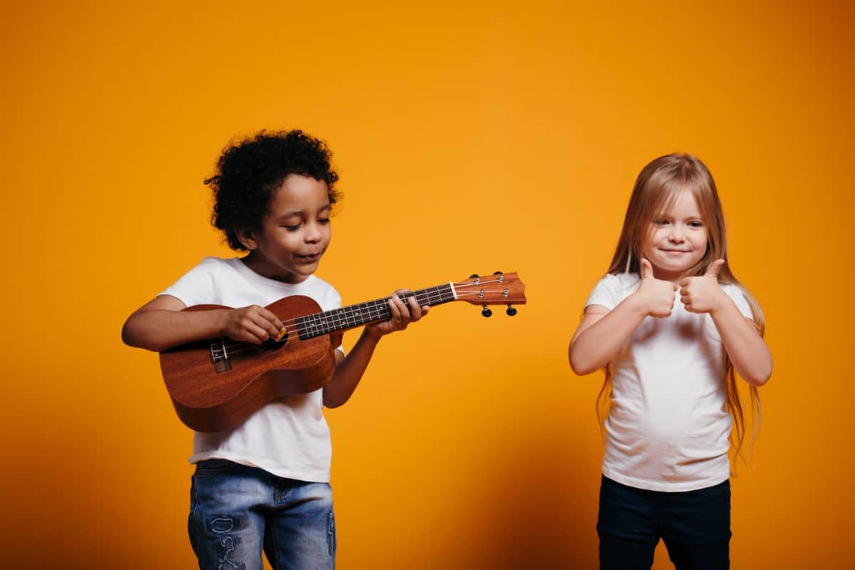 A boy and a girl stand next to each other on a yellow background. The boy has darker skin and is holding a small guitar