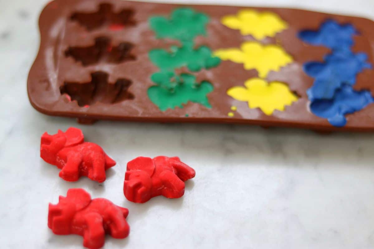 3 red dinosaur crayons sit in front of a brown silicone tray filled with lines of green, yellow and blue dinosaurs