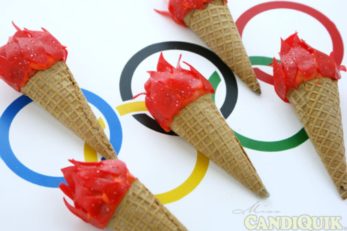 on a cloth of olympic rings are 5 ice cream cones made to look like olympic torches