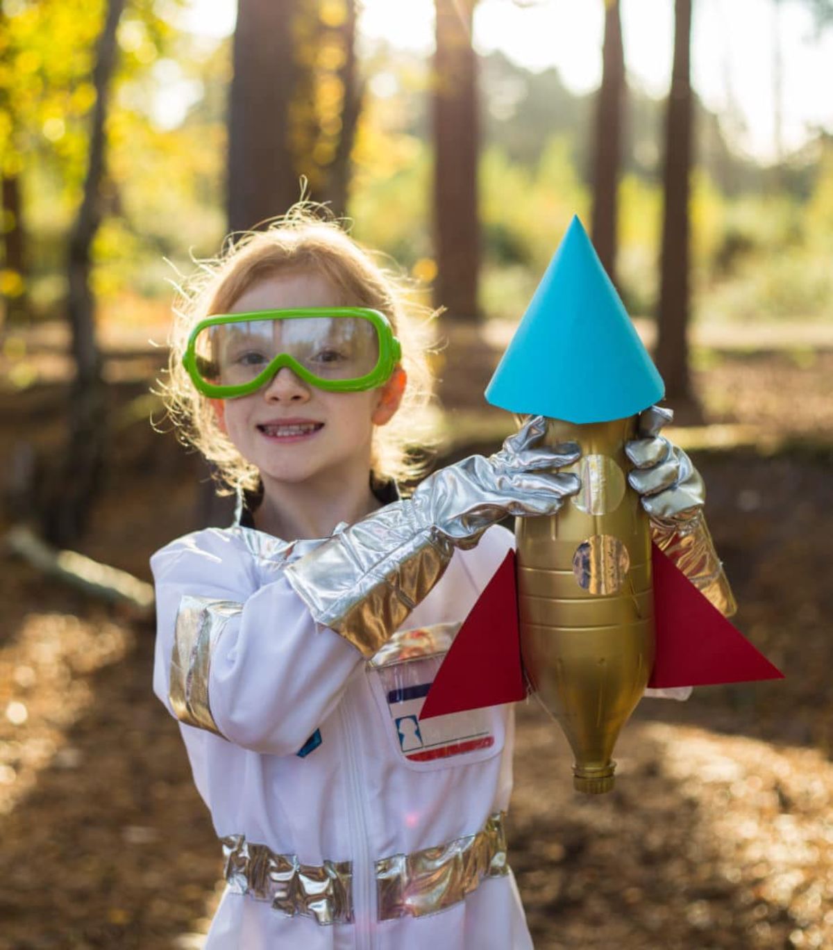 A girl dressed as an astronaut wearing goggles and holds uo a rocket made of a water bottle and cardboard