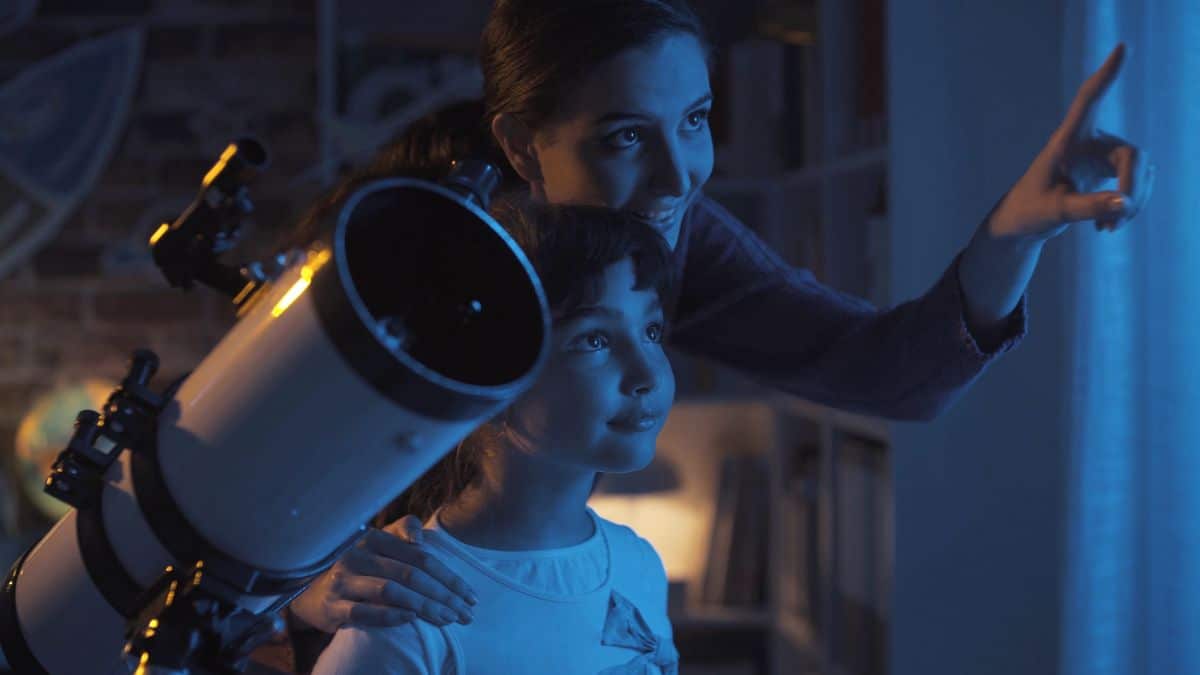a boy and his omther stand next to a telescope with the mother pointing out of the window and up at the sky