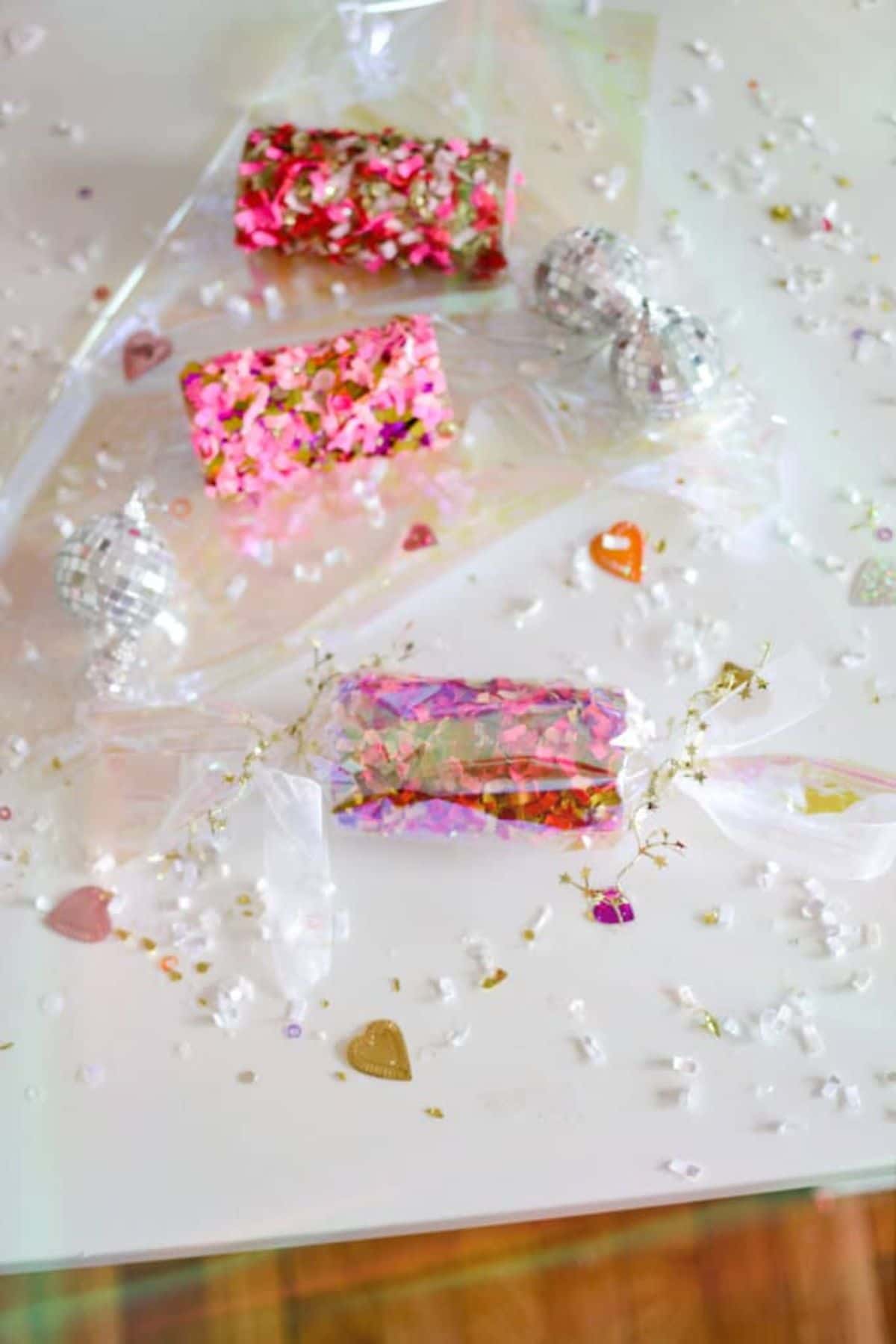 on a white tablee is some clear plastic with glitter bars on it. Glitter and small hearts are scattered around, and one wrapper glitter bar is at the front of the picture