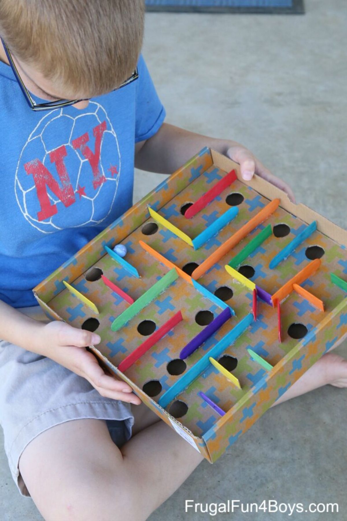 a boy wearing glasses, shorts and a t-shirt holds a cardboard maze, guiding a marble around the walls