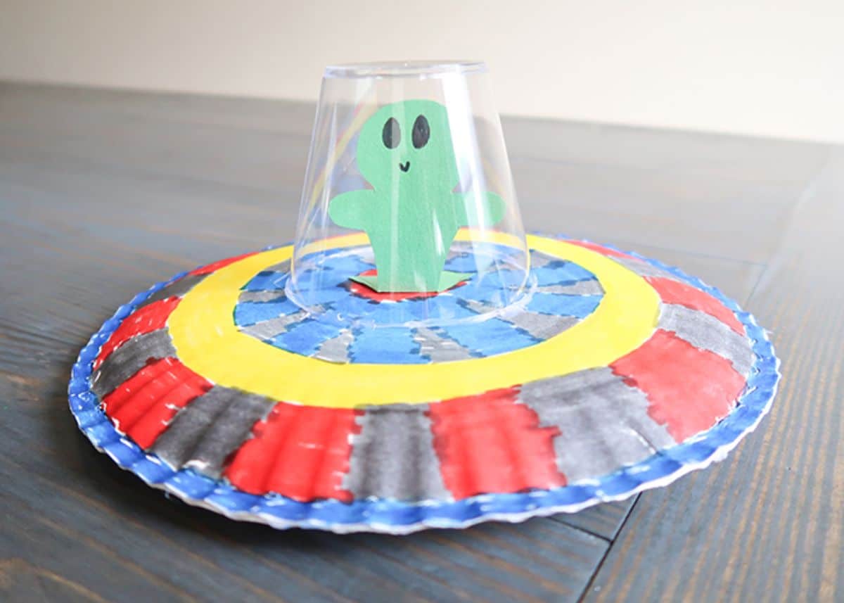 a flying saucer is made out of two paper plates. A paper alien sits inside a clear plastic cup on top of the saucer