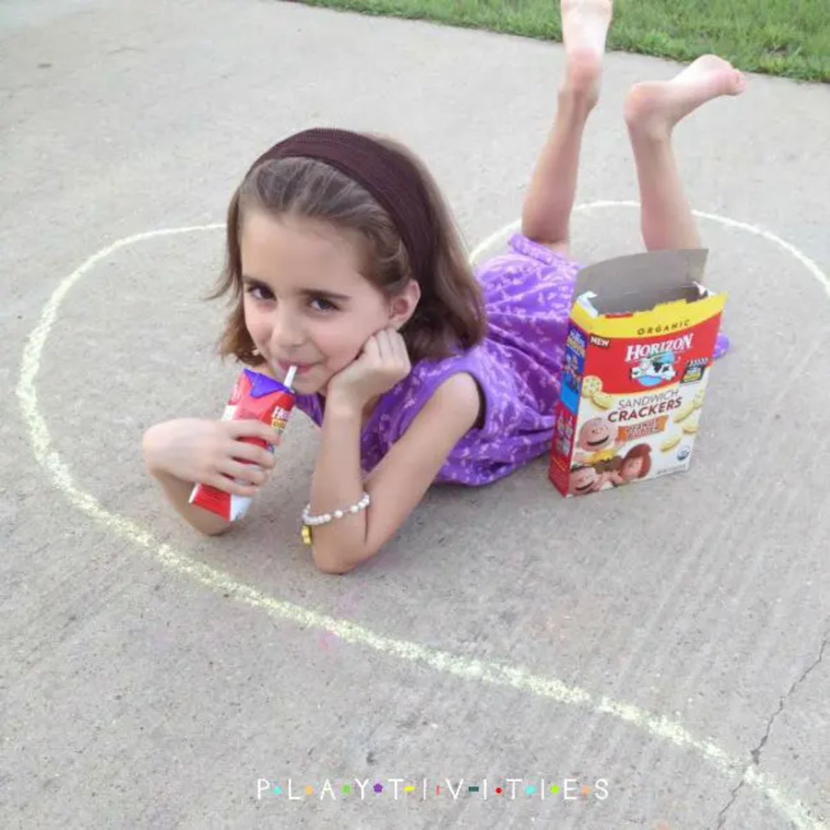 a girl in a purple dress lays on her front on the sidewalk drinking from a juice box through a straw. A box of crackers sits by her side and a chalk outline surrounds her