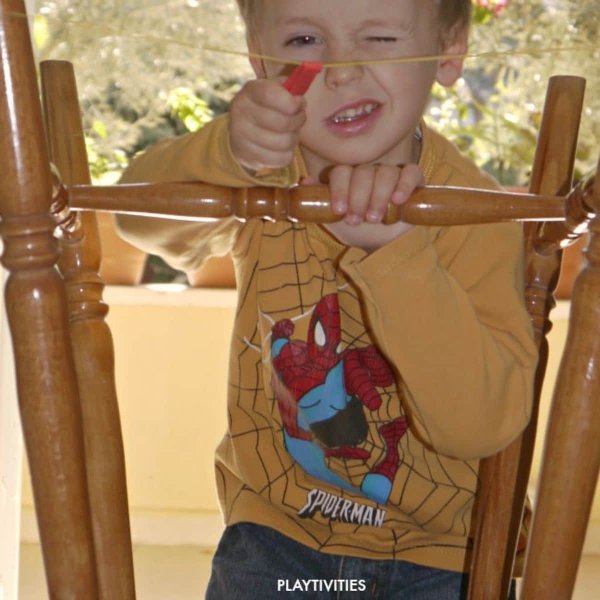a boy in a yellow spiderman shirt sits behind the legs of a chair. He is pulling back a slingshot and squinting at the camera