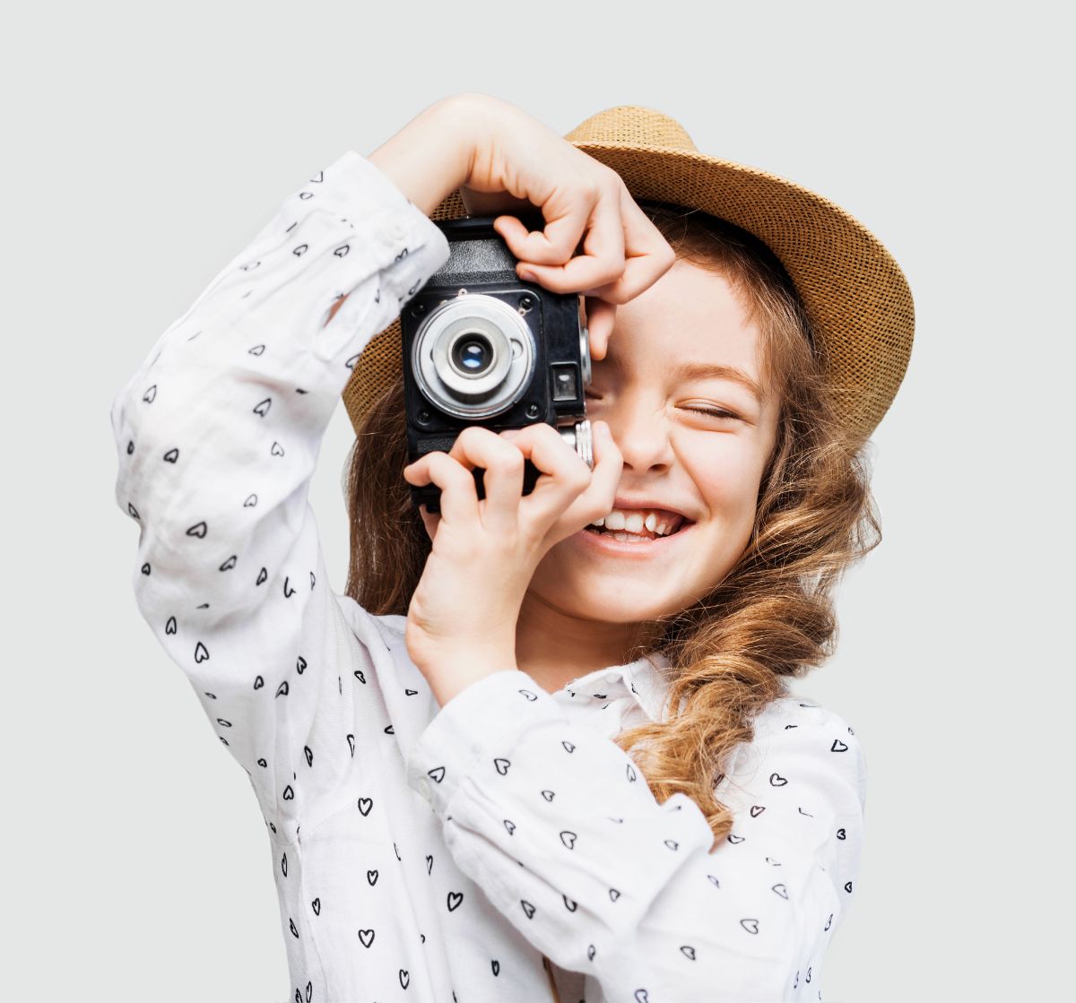 a girl in a white shirt with hearts and a straw hat faces the camera holding a camera up her face as if taking a photo