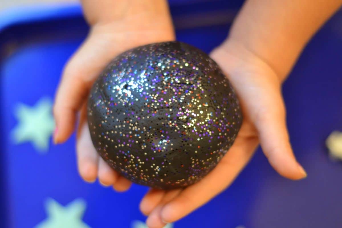 two child's hands hold a black ball of playdough studded with glitter