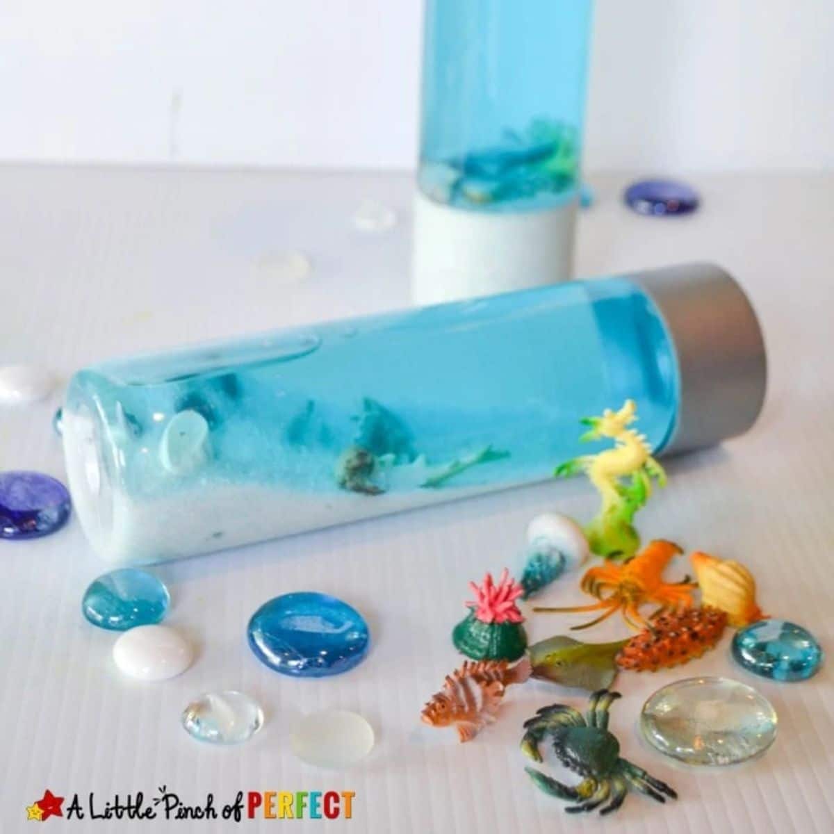 a water bottle is laid down filled with white sand, blue water and toy sea creatures. Other sea creatures are scattered in front of it