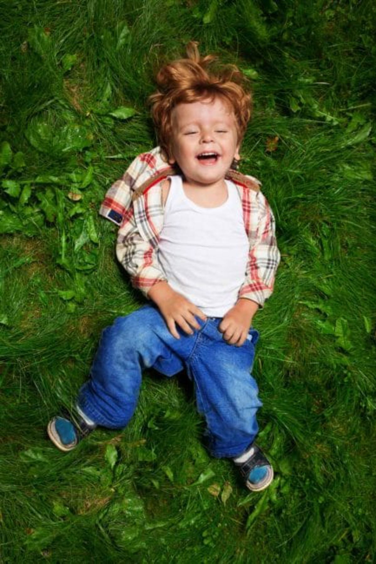 a young boy with red hair, blue jeans and a checked red and white shirt lays on some grass, looking u pat the camera and laughing