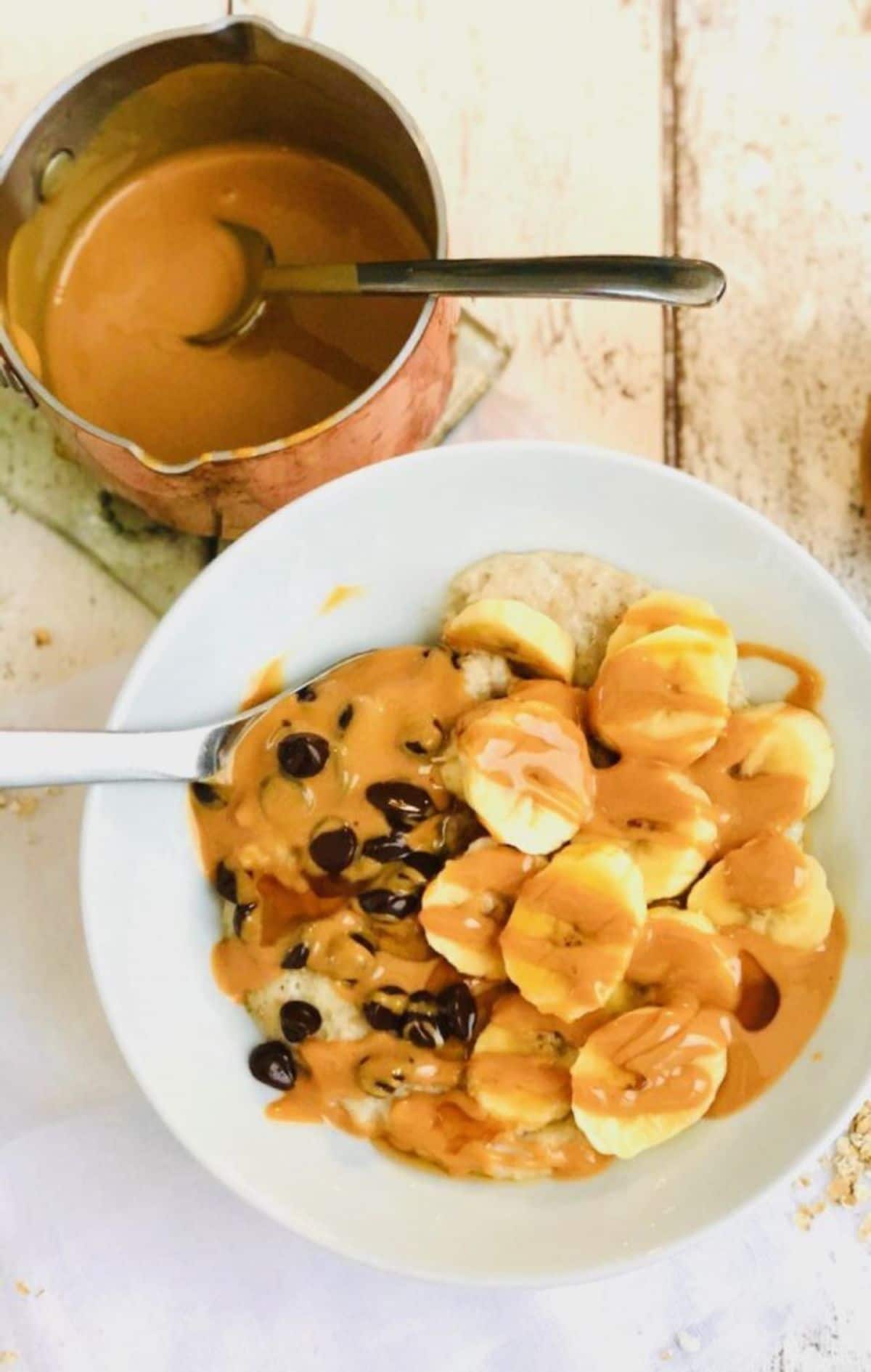 a whote bowl filled with porridge covered in banana slices, raisins, and sauces. A pan of sauce with a spoon in it sits behind