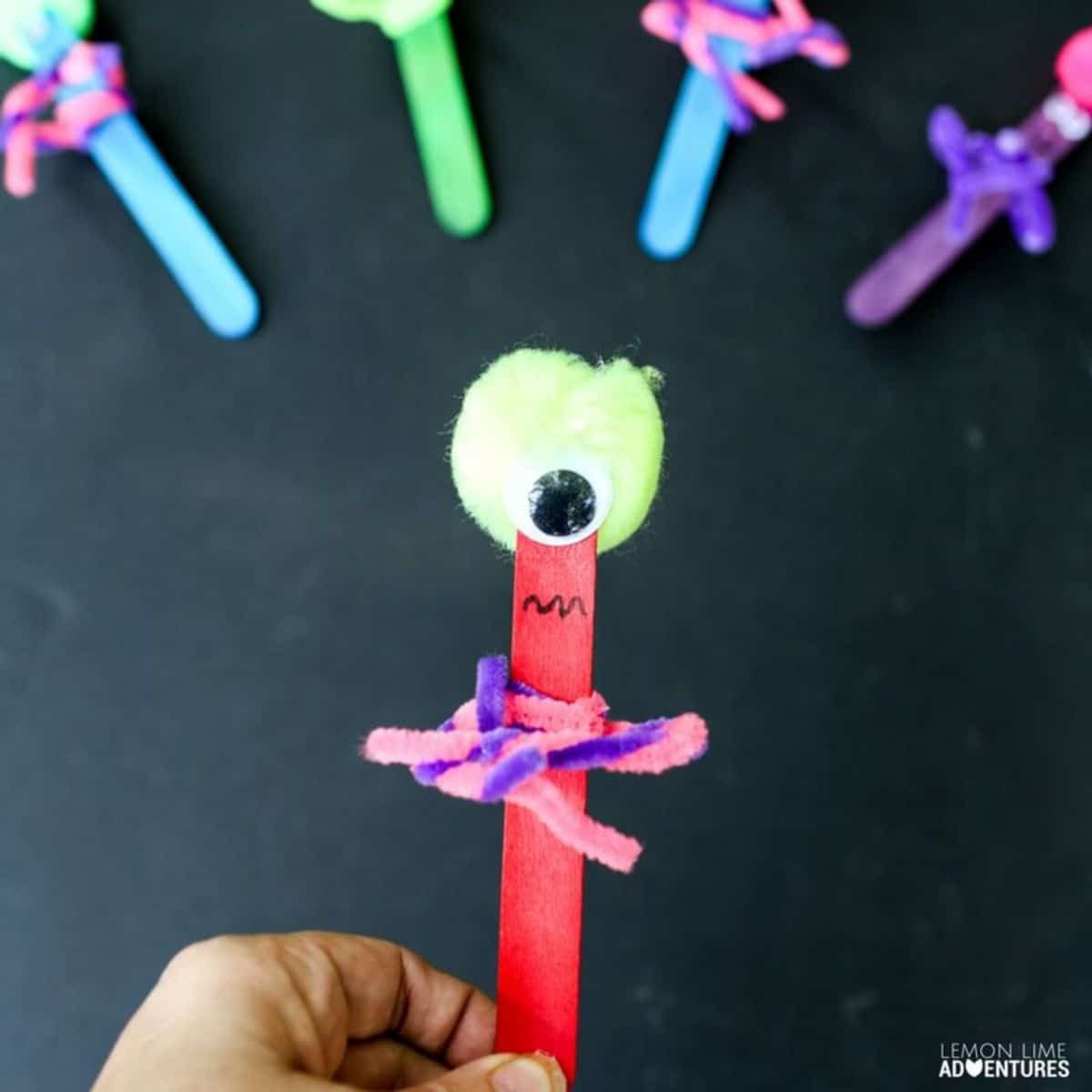 a monster made out of a red popsicle stick, a green pom pom and some pipe cleaners can be seen in front of 4 other creatures