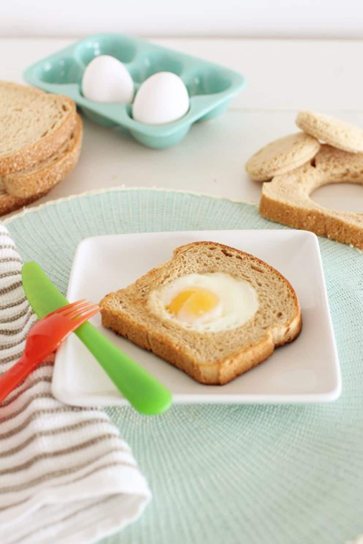 a slice of taost with a fried egg in the middle sits on a square plate. 2 eggs are in an egg box behind, with slices of bread and some plastic cutlery
