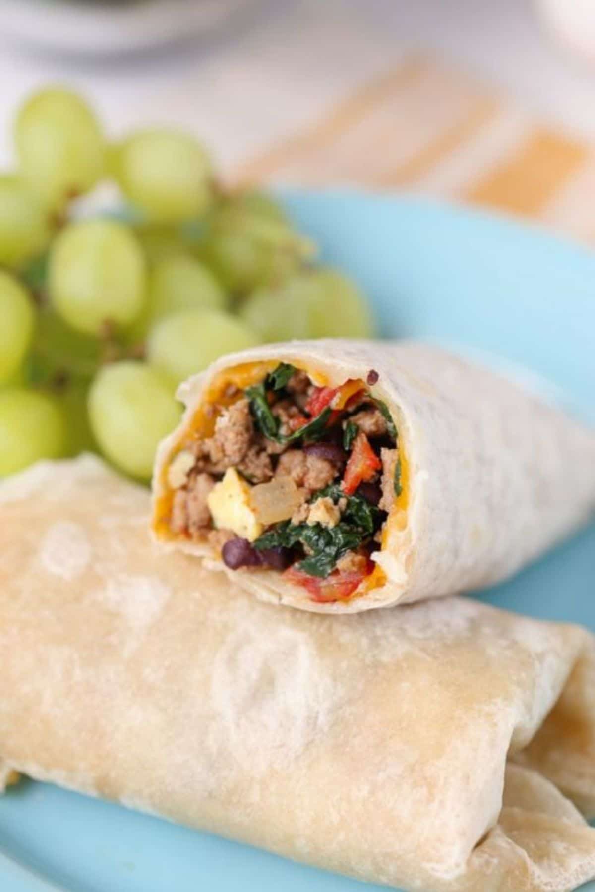 2 tortilla wraps are on a plate filled with sausage, egg and spinach. Behind is a bunch of grapes