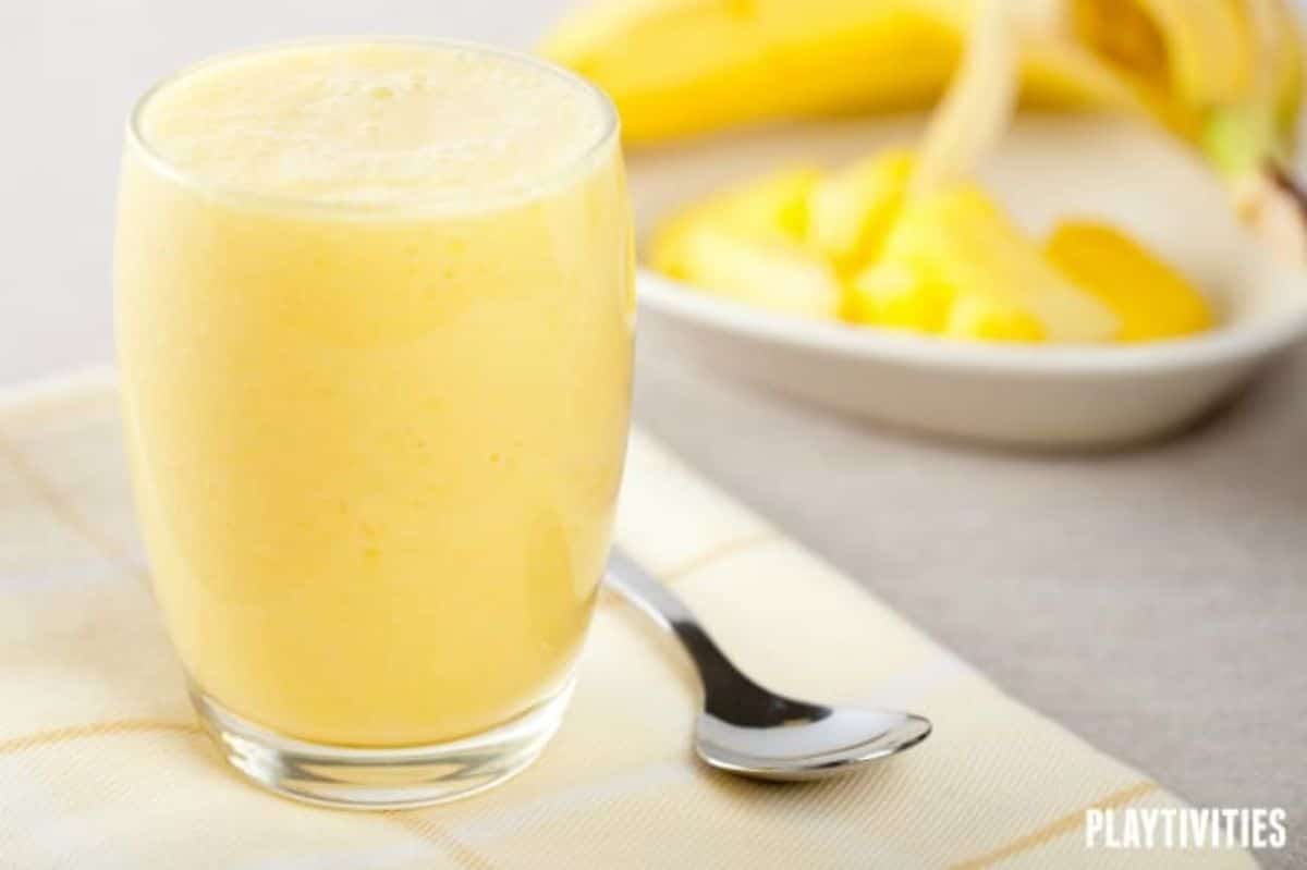 a yellow smoothie fills a glass with a bowl of pineapple and a peeled banana is behind it