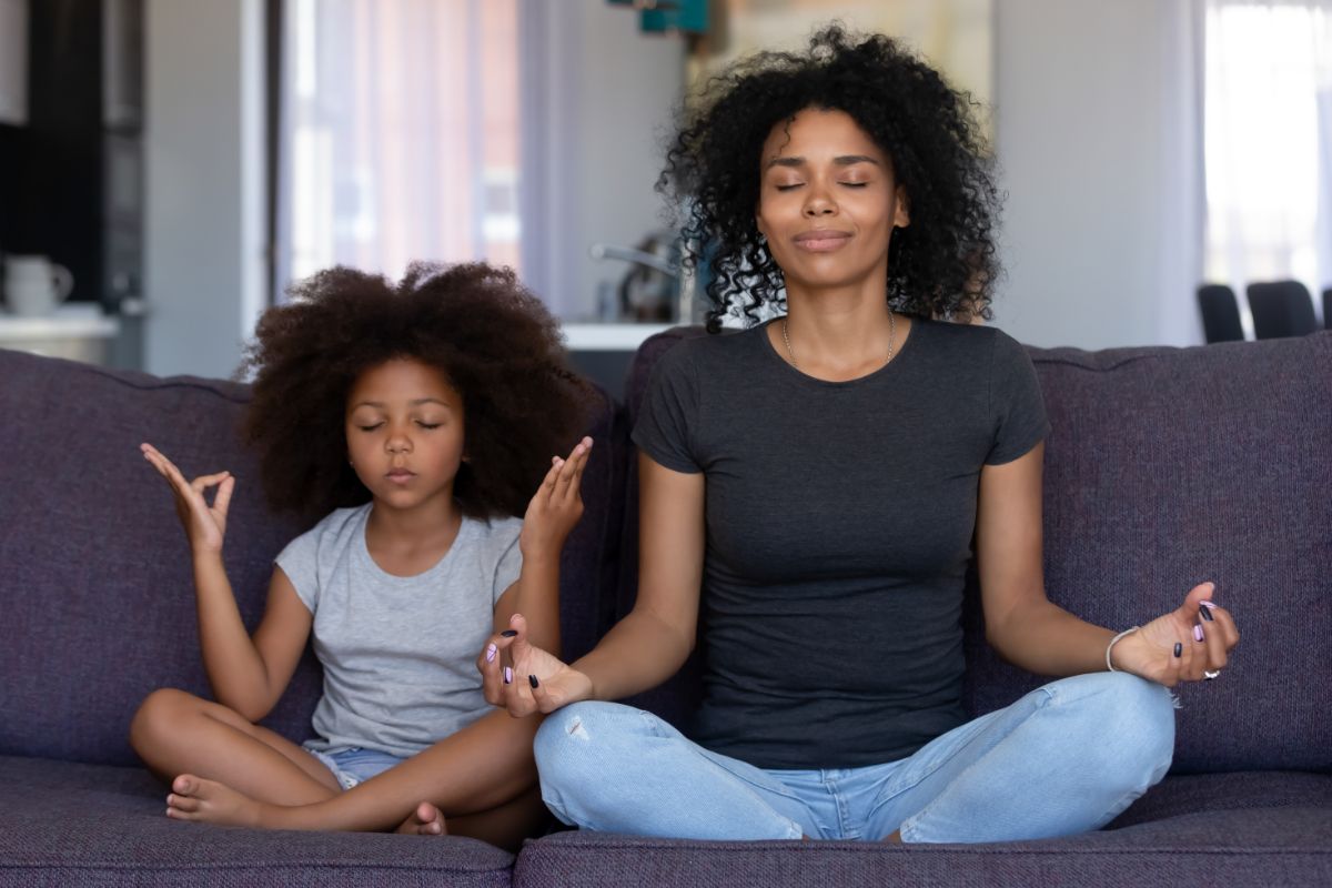 a black mother and daughter sit on a sofa cross-legged, meditating
