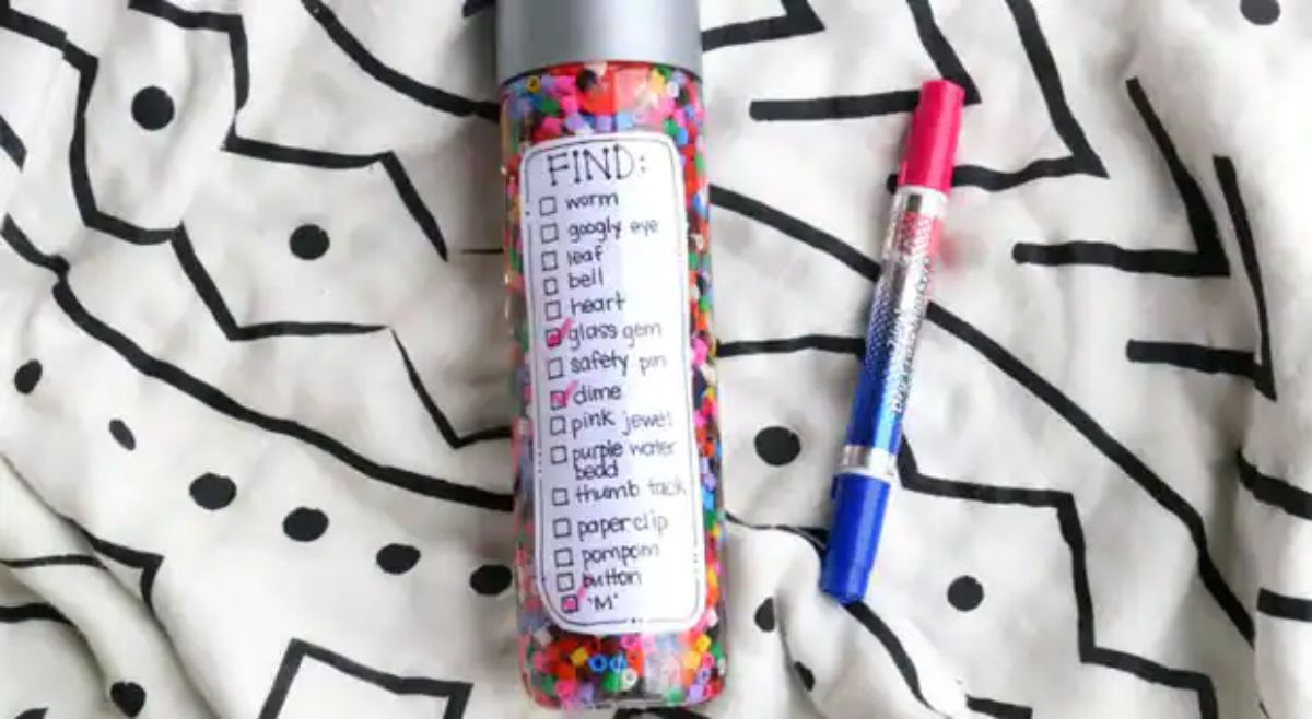 on a black and white patterned sheet is a drinks bottle filled with hama beads. It is labelled with "FIND:" and a list of items. Beside it is a pen