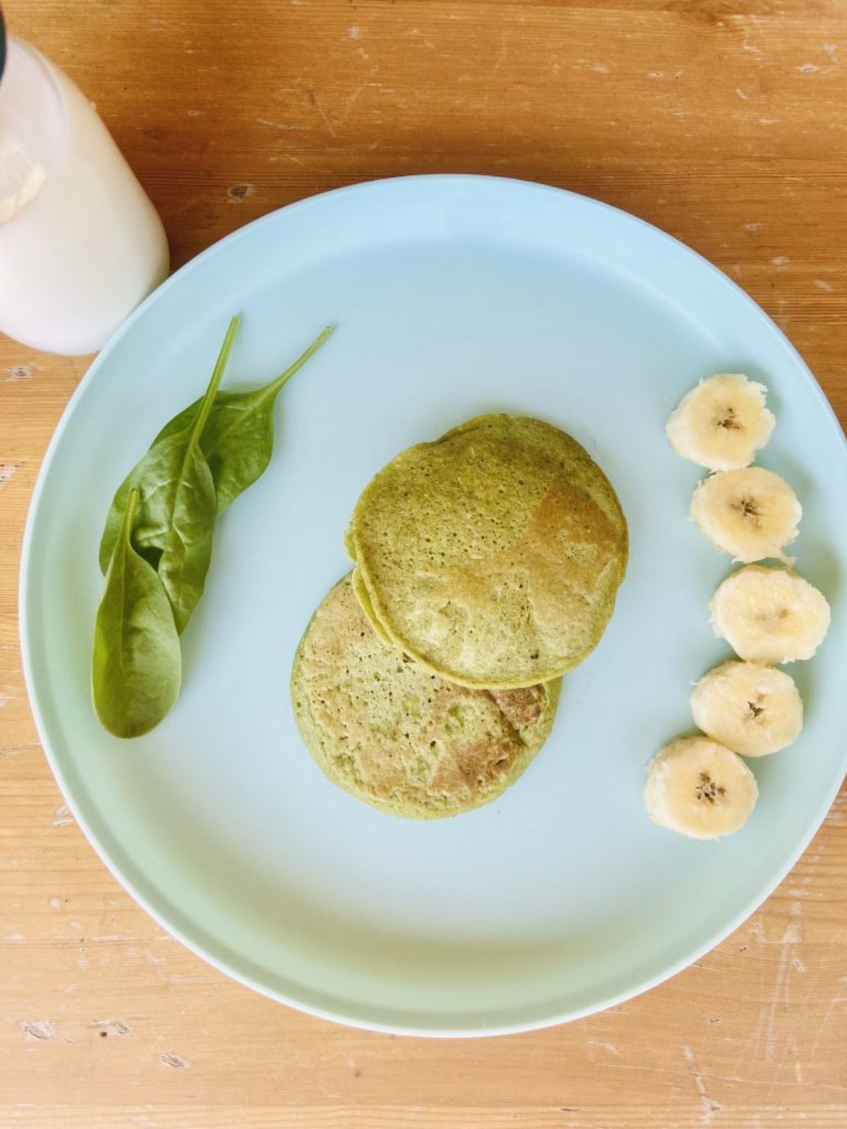 a white plate has spinach leaves on one side, banana slices on the otehr and 2 spinach pancakes in the middle