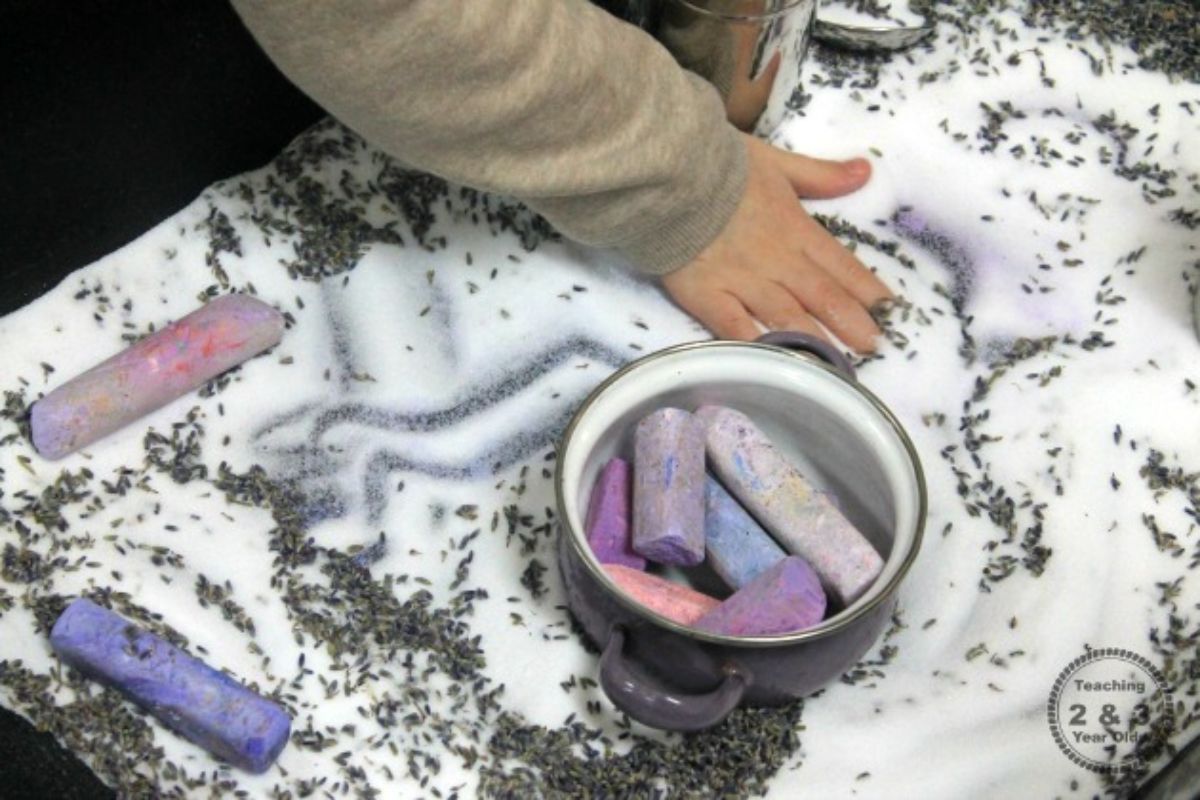 A hand traces patterns in a tray full of salt and lavender. A pot full of purple playdough sticks can be seen in the middle