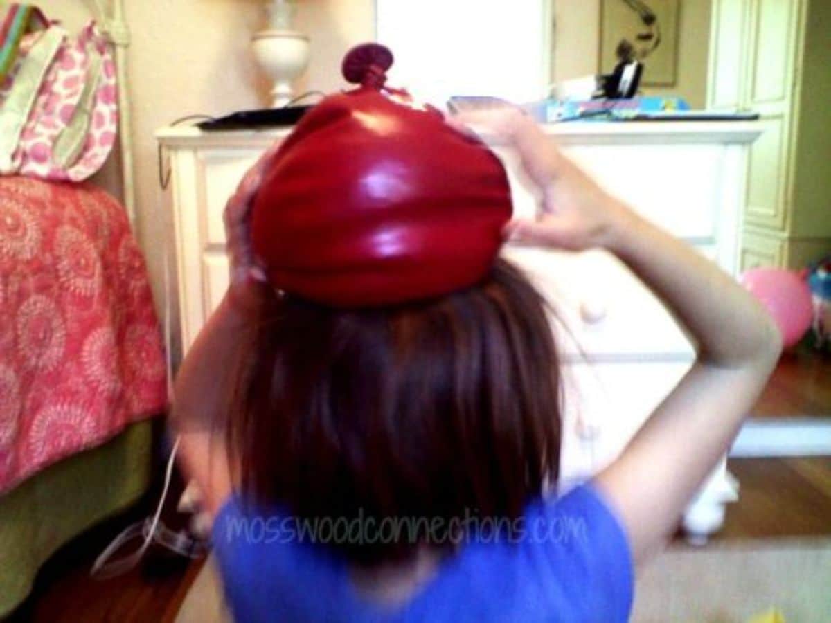 a girl holds a balloon filled with sand on her head, facing away from the camera